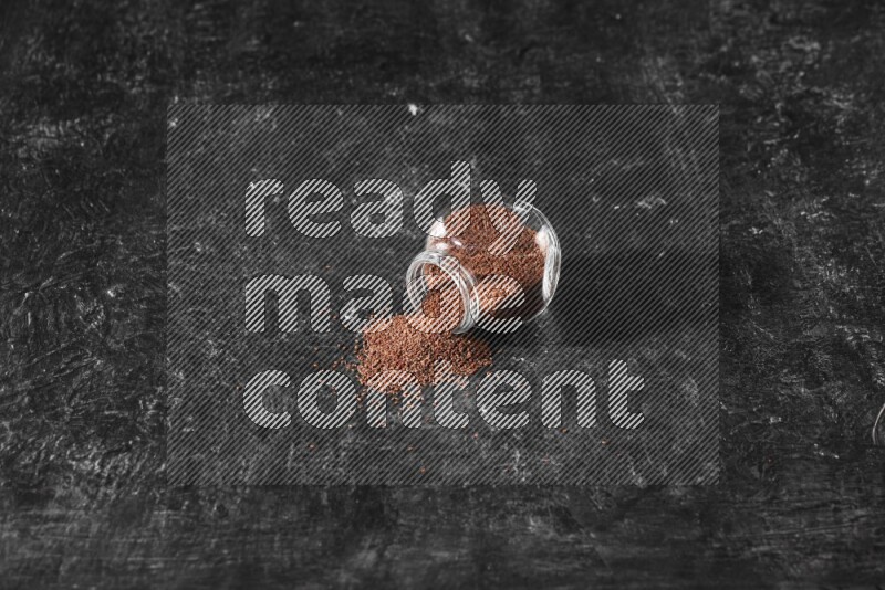 A glass spice jar full of garden cress seeds flipped and seeds spread out a textured black flooring