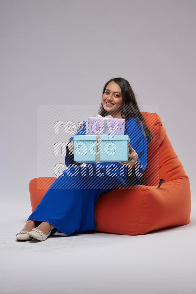 A woman sitting on an orange beanbag wearing Jalabeya holding a gift box