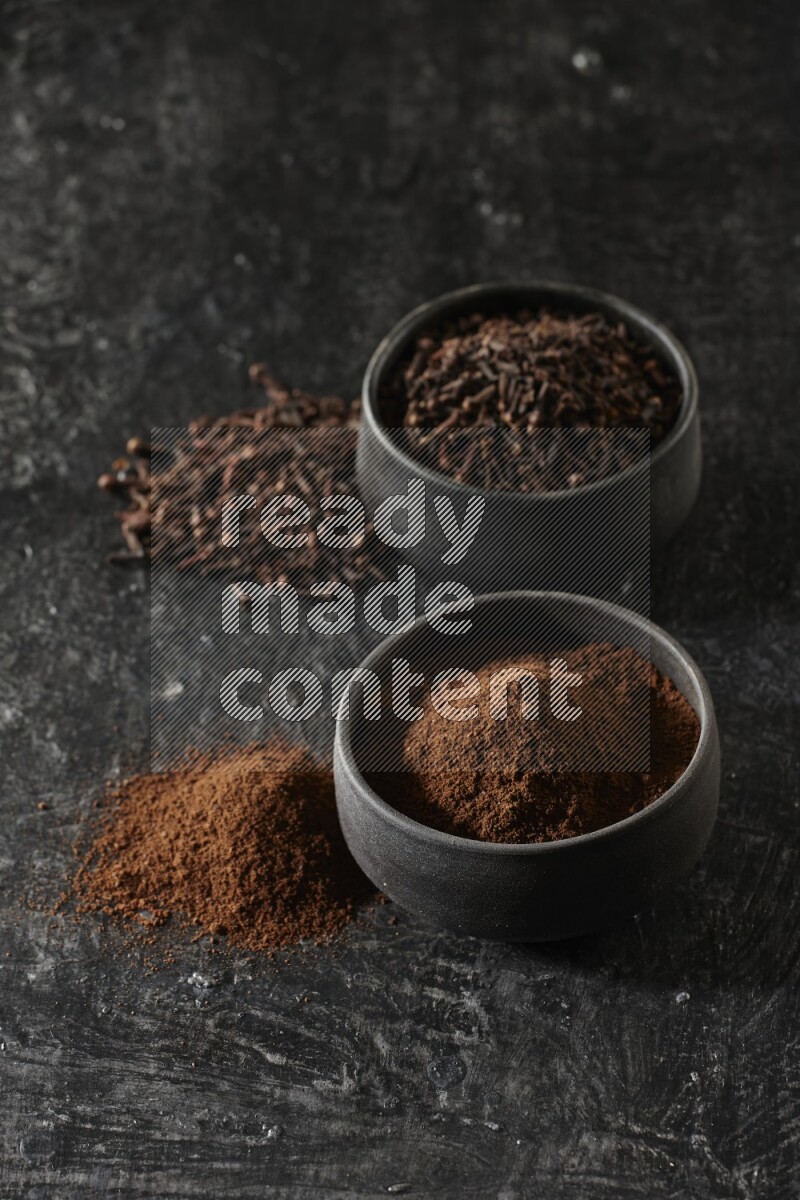 2 Black pottery bowls full of cloves and the other full of cloves powder on textured black flooring