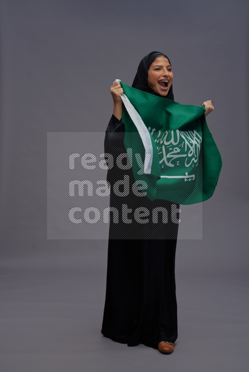 Saudi woman wearing Abaya standing holding Saudi flag on gray background