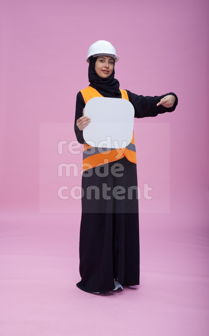 Saudi woman wearing Abaya with engineer vest and helmet standing holding social media sign on pink background