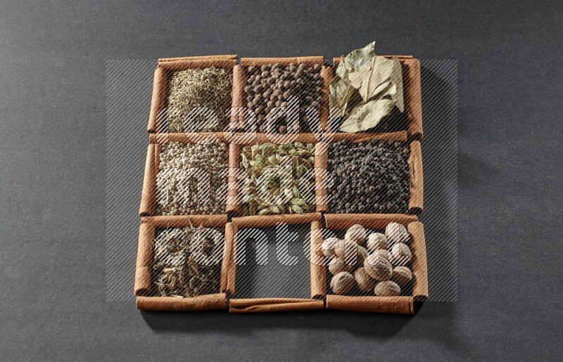 9 squares of cinnamon sticks full of cardamom in the middle surrounded by nutmeg, black pepper, bay laurel leaves, allspice, cumin, white pepper, dried basil and salt on black flooring