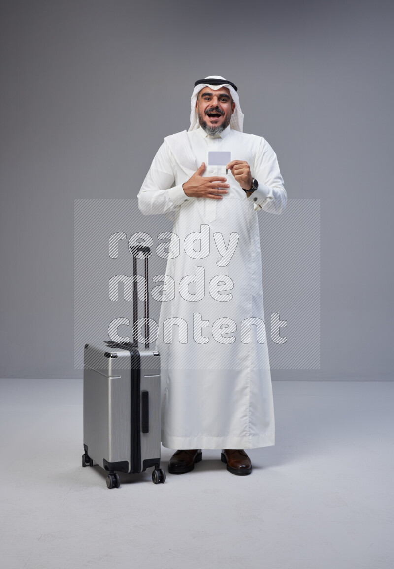 Saudi man wearing Thob and white Shomag standing holding Travel bag and ATM card on Gray background