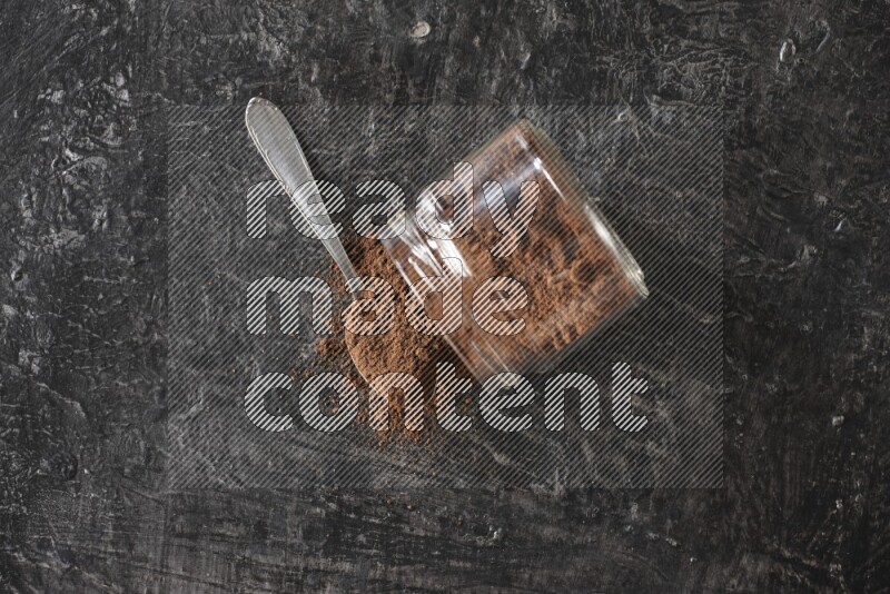 A flipped glass jar and metal spoon full of cloves powder with cloves spread on a textured black flooring