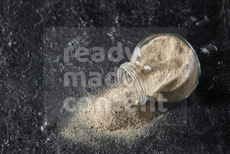 A flipped herbal glass jar full of white pepper powder with spilled powder on textured black flooring