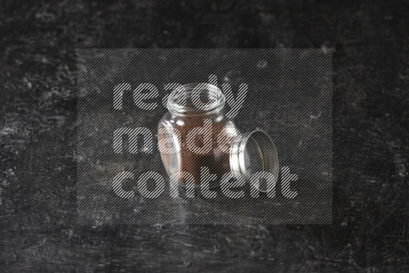 A glass spice jar full of cloves powder on textured black flooring