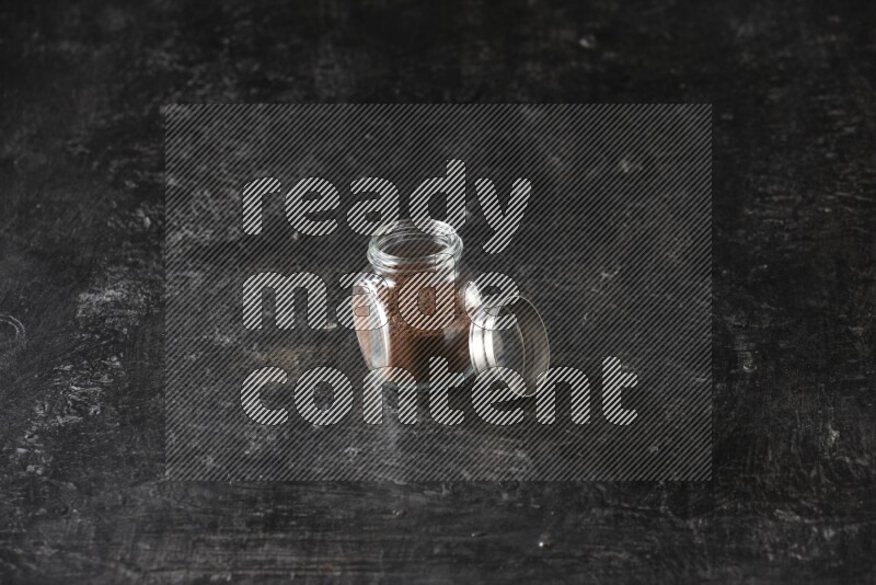 A glass spice jar full of cloves powder on textured black flooring