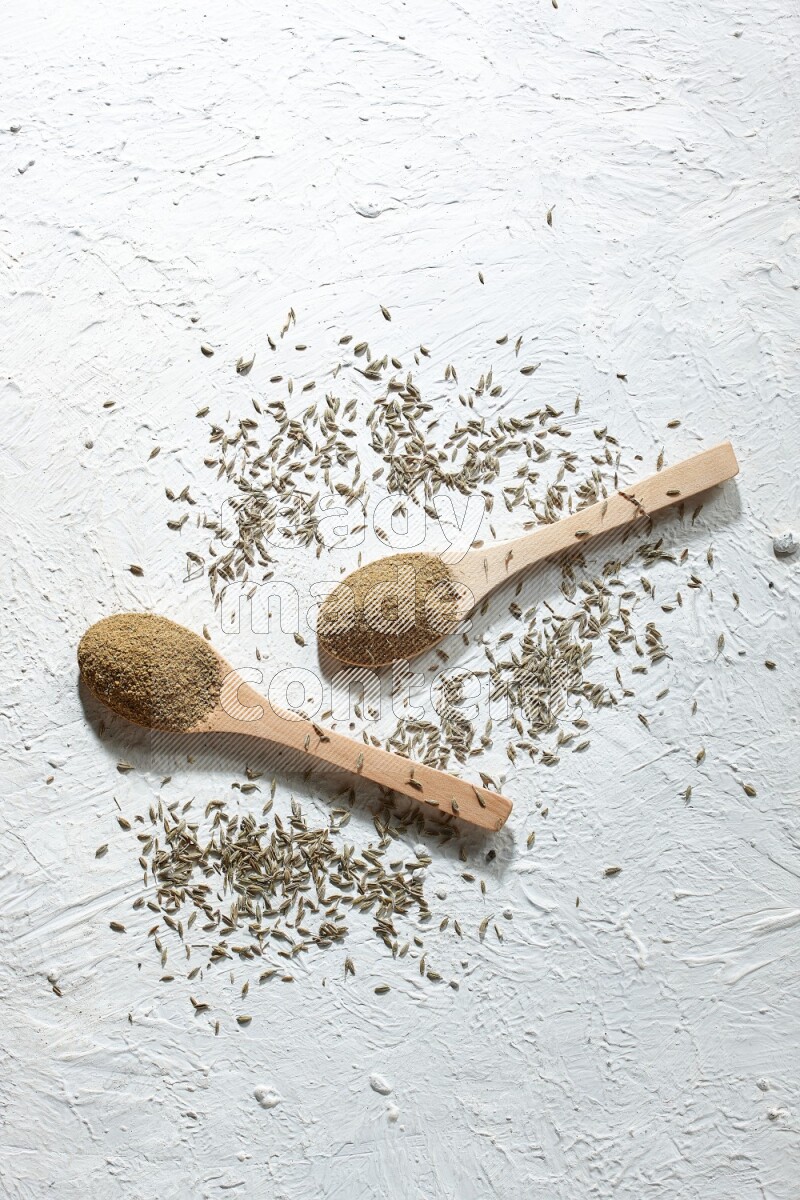 2 Wooden spoons full of cumin powder and cumin seeds on textured white flooring