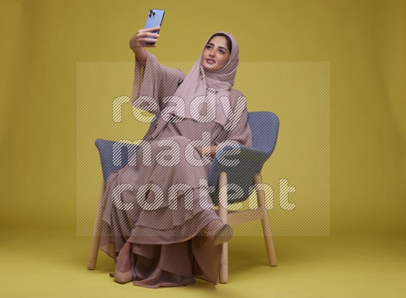 A woman sitting on a blue chair and Taking a Selfie  Setting  on a Yellow Background wearing Brown Abaya with Hijab