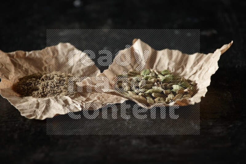 Cardamom seeds and cardamom powder in 2 crumpled pieces of paper on textured black flooring