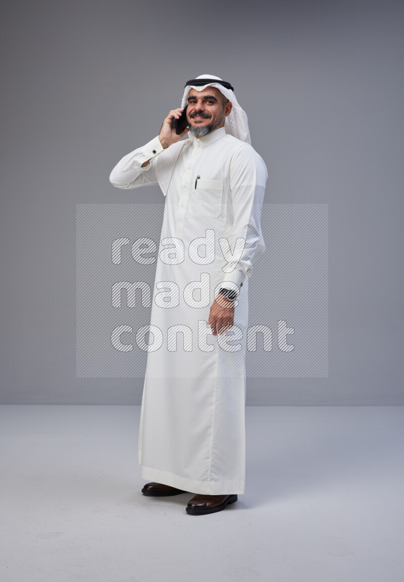 Saudi man Wearing Thob and white Shomag standing talking on phone on Gray background