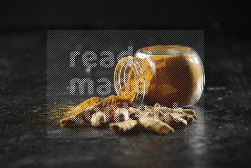 A flipped glass spice jar full of turmeric powder and powder spilled out of it with dried whole fingers on textured black flooring
