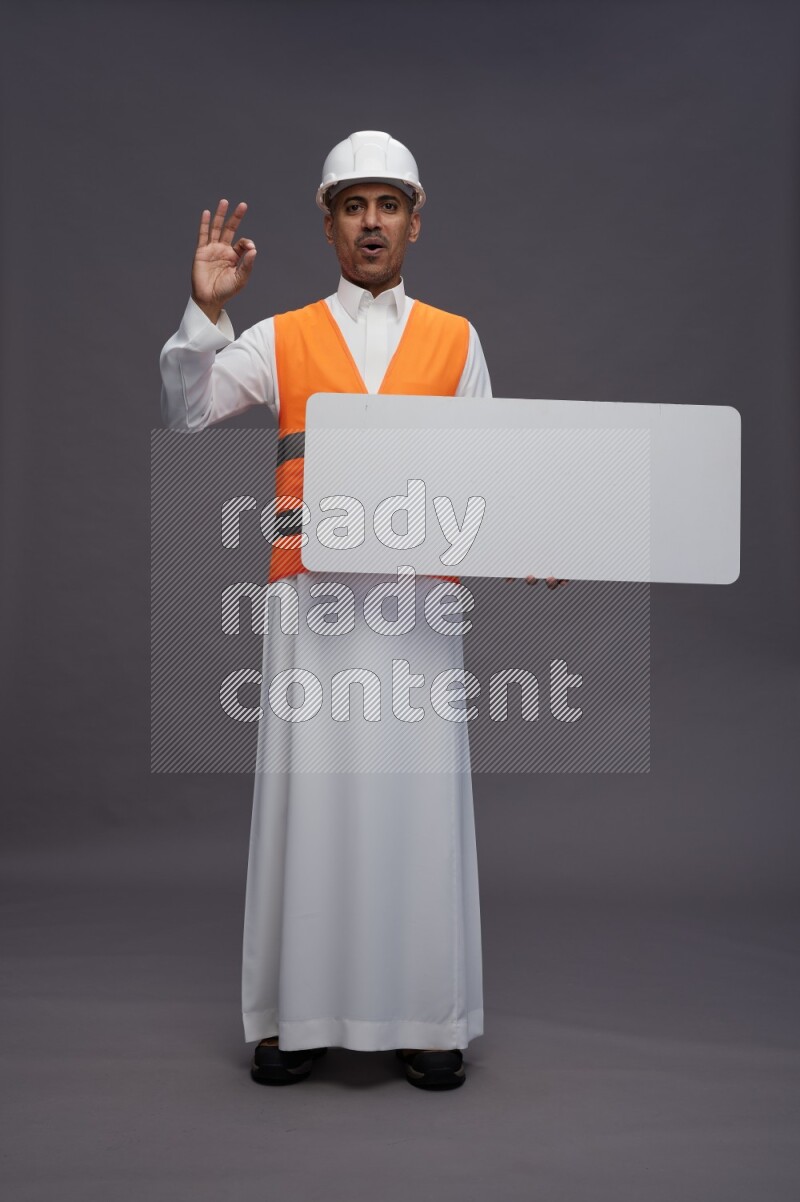 Saudi man wearing thob with engineer vest standing holding board on gray background
