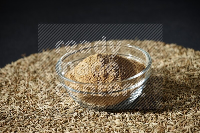 A glass bowl full of cumin powder surrounded by cumin seeds on black flooring