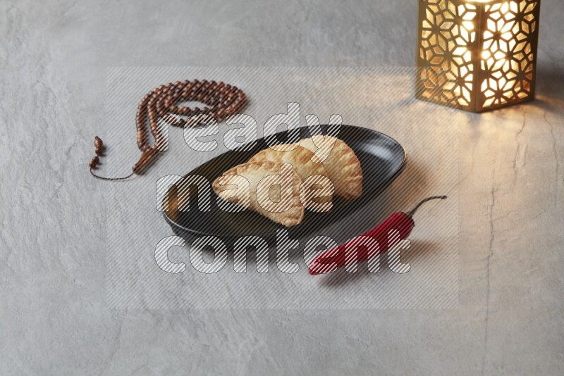 Three fried sambosas in an oval shaped black plate and a red sauce in a black round ramekin with a brown misbaha and a golden lantern on the side on a gray background