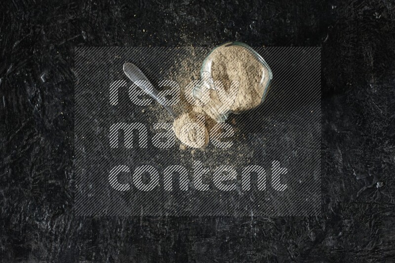 وعاء زجاجي للتوابل مقلوب وملعقة معدنية ممتلئان ببودرة الهيل والبودرة تناثرت منه على خلفية سوداء