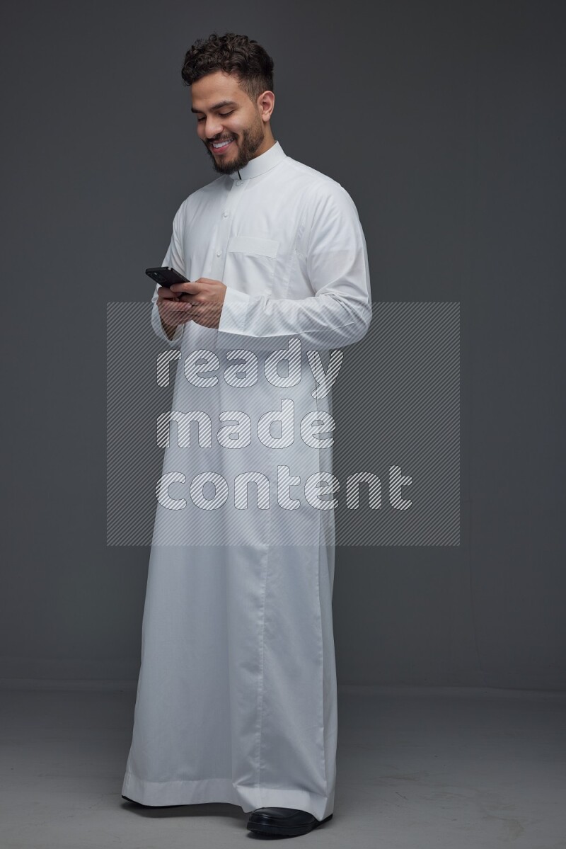 A Saudi man wearing Thobe standing and using his phone eye level on a gray background