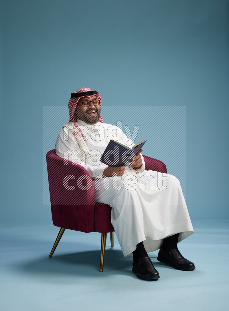 Saudi Man with shimag sitting on chair reading book on blue background