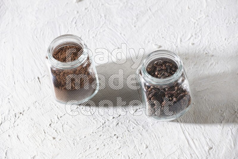2 glass jars full of cloves powder and cloves grains on a textured white flooring