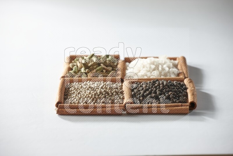 4 مربعات من أعواد القرفة ممتلئة بالهيل والملح والفلفل الأبيض والفلفل الأسود على أرضية بيضاء