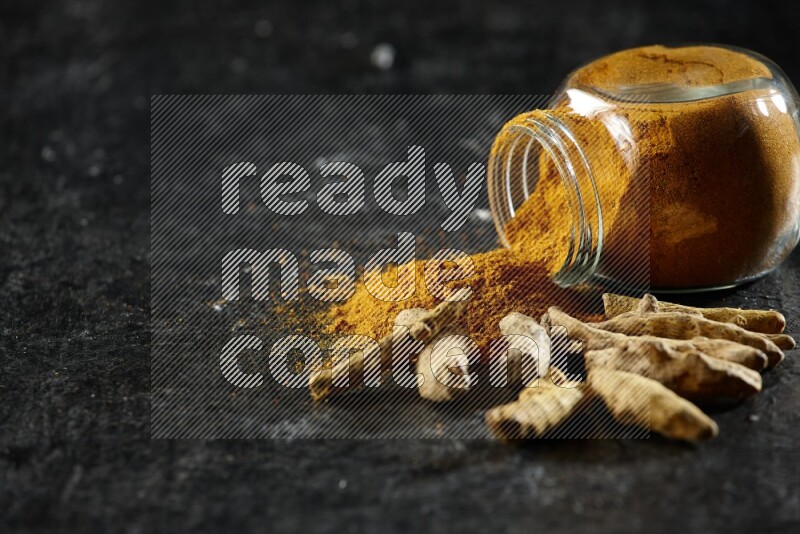 A flipped glass spice jar full of turmeric powder and powder spilled out of it with dried whole fingers on textured black flooring