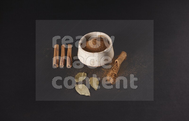 Cinnamon powder in a white pottery bowl and cinnamon sticks and laurel leaves on black background