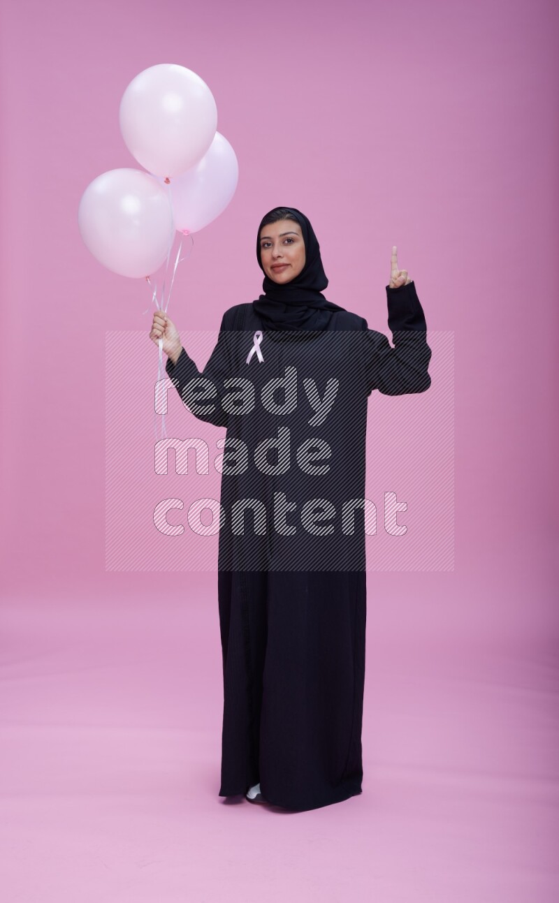 Saudi woman wearing pink ribbon on Abaya standing holding balloons on plain pink background