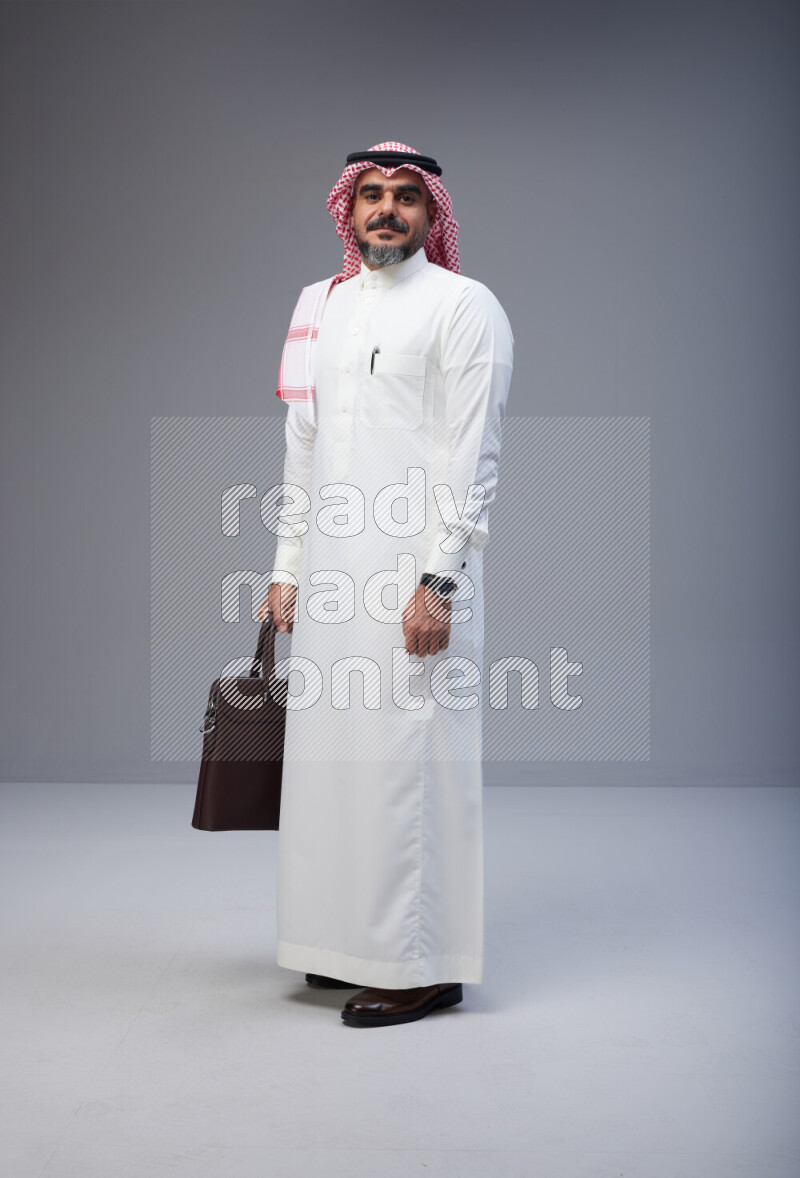 Saudi man Wearing Thob and red Shomag standing holding bag on Gray background
