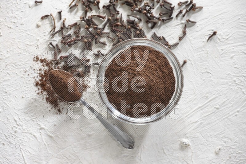 A glass bowl and a metal spoon full of cloves powder with cloves grains spread on a textured white flooring
