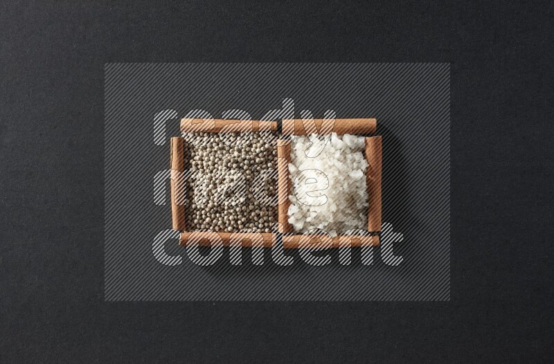 مربعان من أعواد القرفة ممتلئان بحبوب الفلفل الأسود والملح علي خلفية سوداء