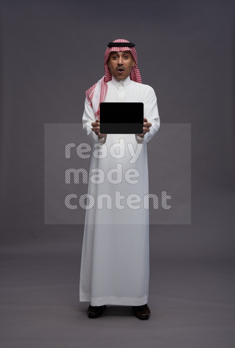 Saudi man wearing thob and shomag standing showing tablet to camera on gray background