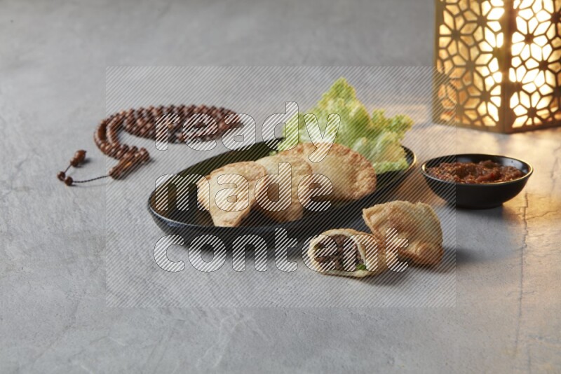 Three fried sambosas in an oval shaped black plate and a red sauce in a black round ramekin with a brown misbaha and a golden lantern on the side on a gray background
