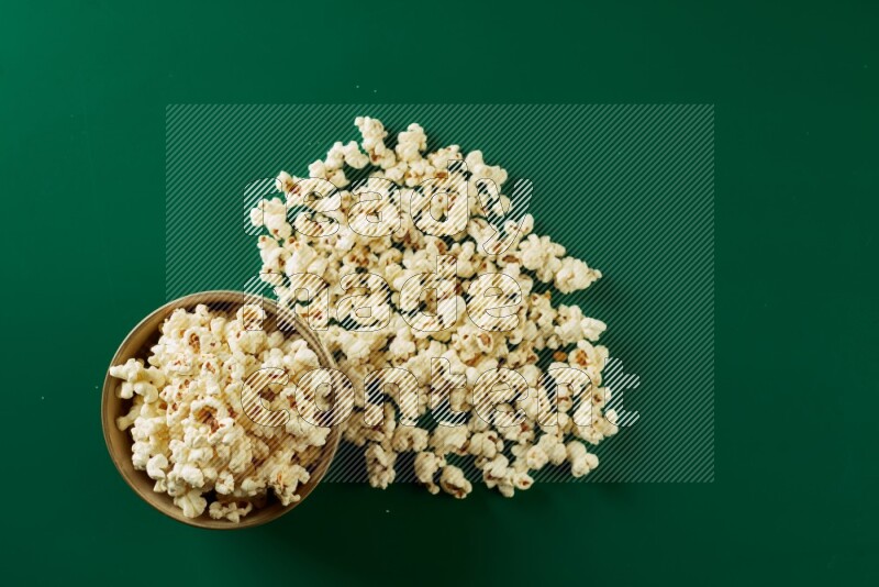 An off white ceramic bowl full of popcorn with popcorn beside it on a green background in a top view shot