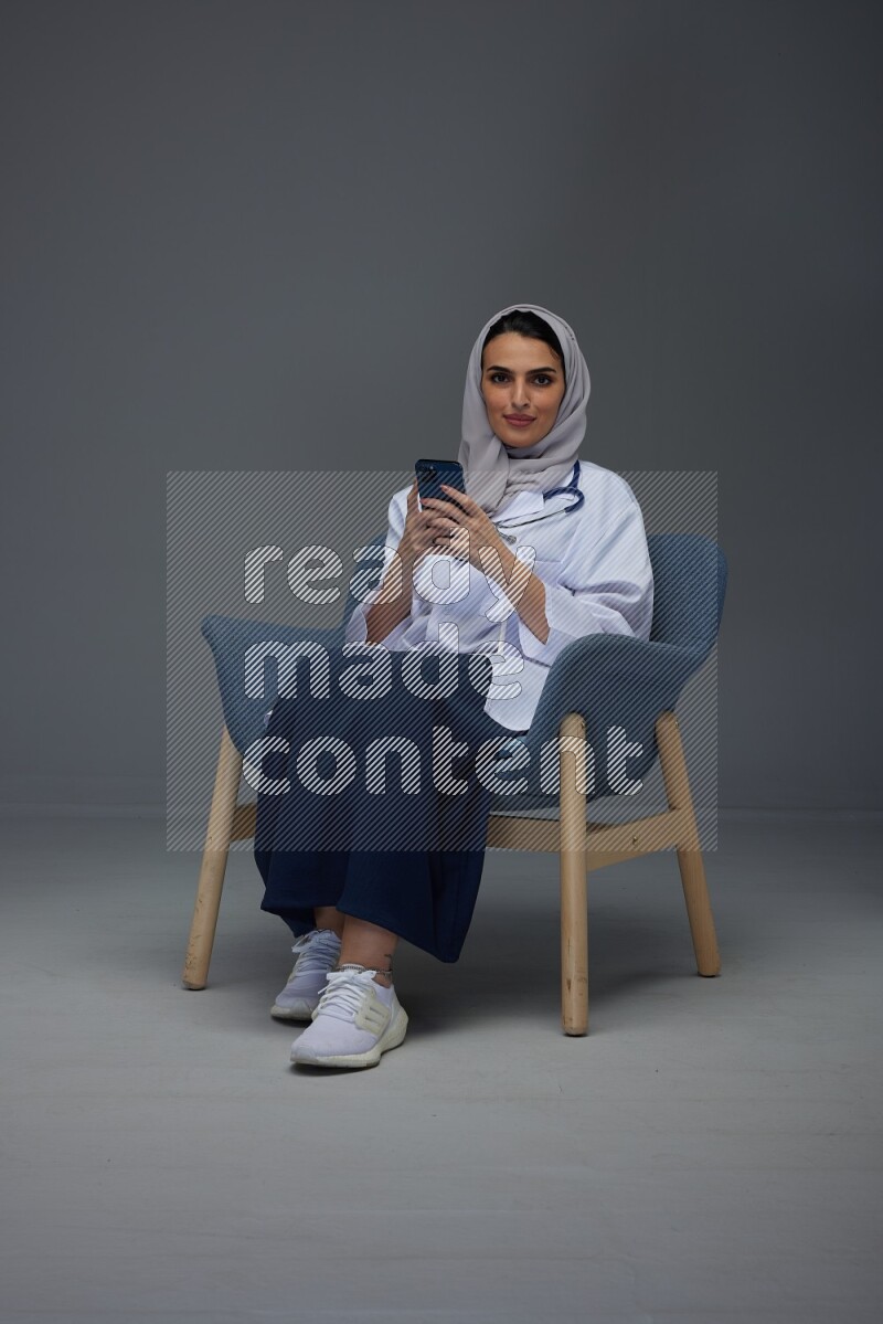 A doctor wearing a light gray head scarf sitting on a dark grey chair and using a laptop while talking in the phone eye level on a grey background