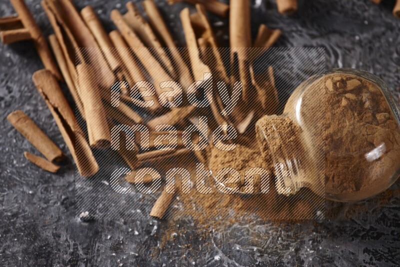 Herbal glass jar full cinnamon powder flipped and a metal spoon full of powder surrounded by cinnamon sticks on textured black background in different angles