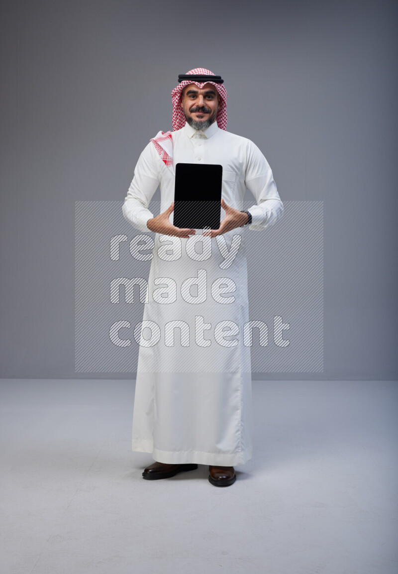 Saudi man Wearing Thob and red Shomag standing showing tablet to camera on Gray background