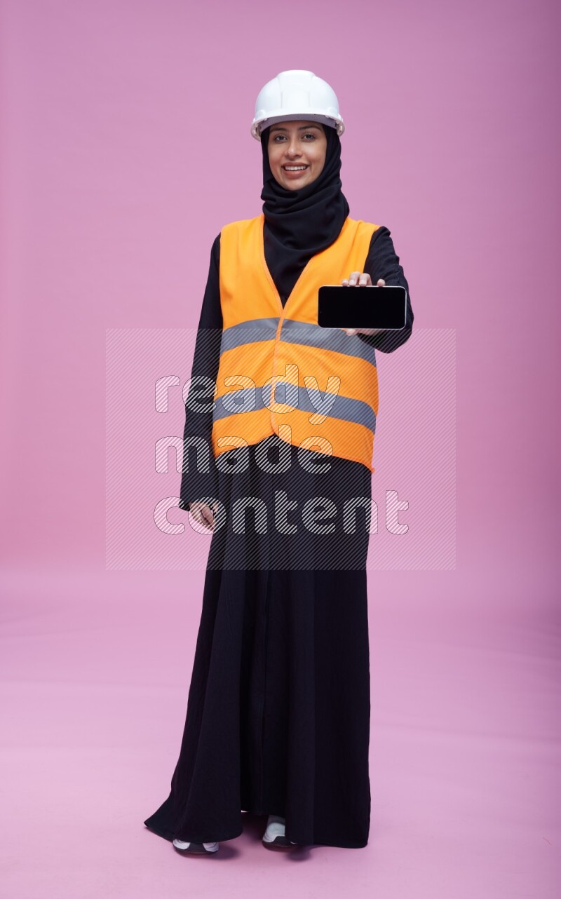 Saudi woman wearing Abaya with engineer vest and helmet standing showing phone to camera on pink background