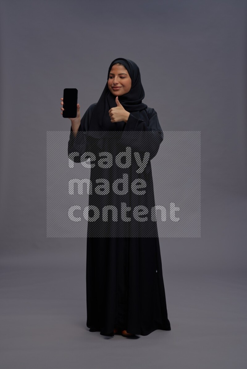 Saudi woman wearing Abaya standing showing phone to camera on gray background