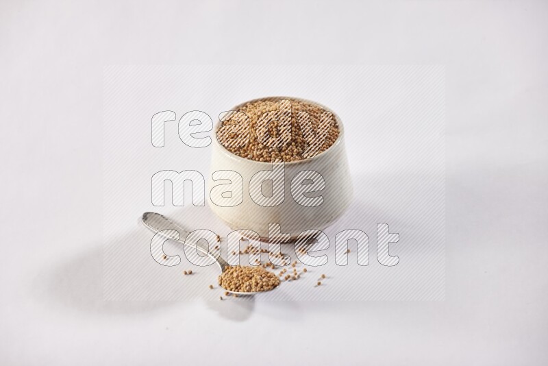 A beige pottery bowl and a metal spoon full of mustard seeds on white flooring