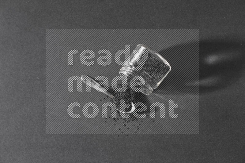 A flipped glass jar full of black seeds and seeds spread out with a metal spoon full of the seeds on a black flooring