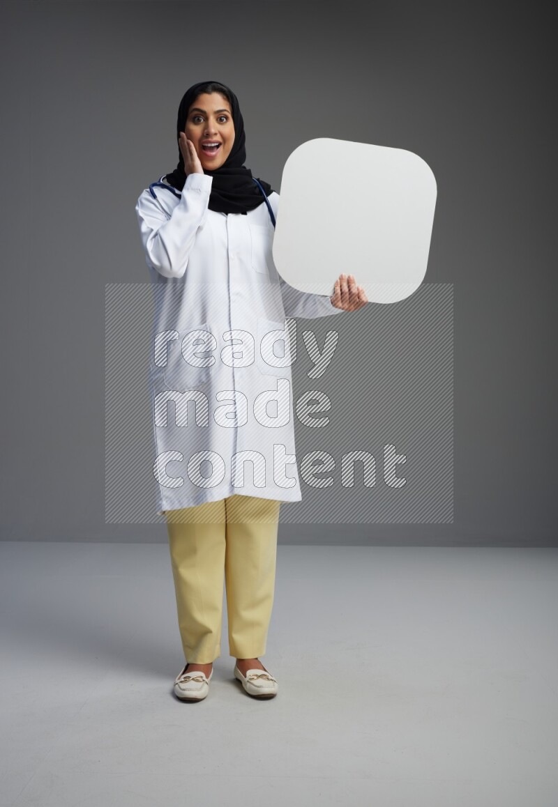 Saudi woman wearing lab coat with stethoscope standing holding social media sign on Gray background