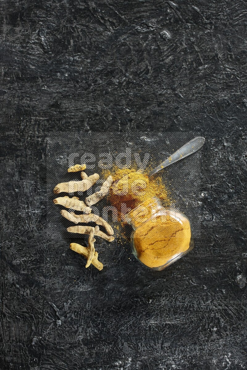 A flipped glass spice jar and metal spoon full of turmeric powder and powder spilled out of it with dried whole fingers on textured black flooring