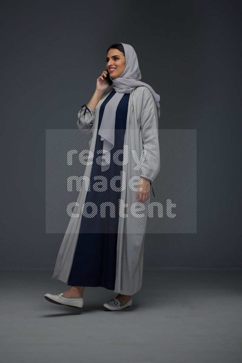 A Saudi woman wearing a light gray Abaya and head scarf standing and talking in the phone eye level on a grey background