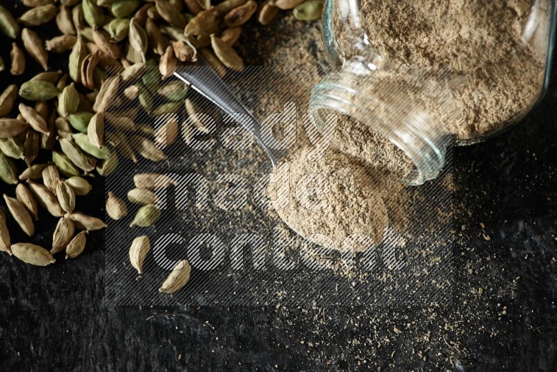 A flipped glass spice jar and a metal spoon full of cardamom powder and cardamom seeds spreaded on textured black flooring