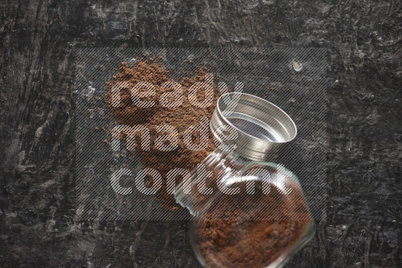 A flipped glass spice jar full of cloves powder and powder came out of it on textured black flooring