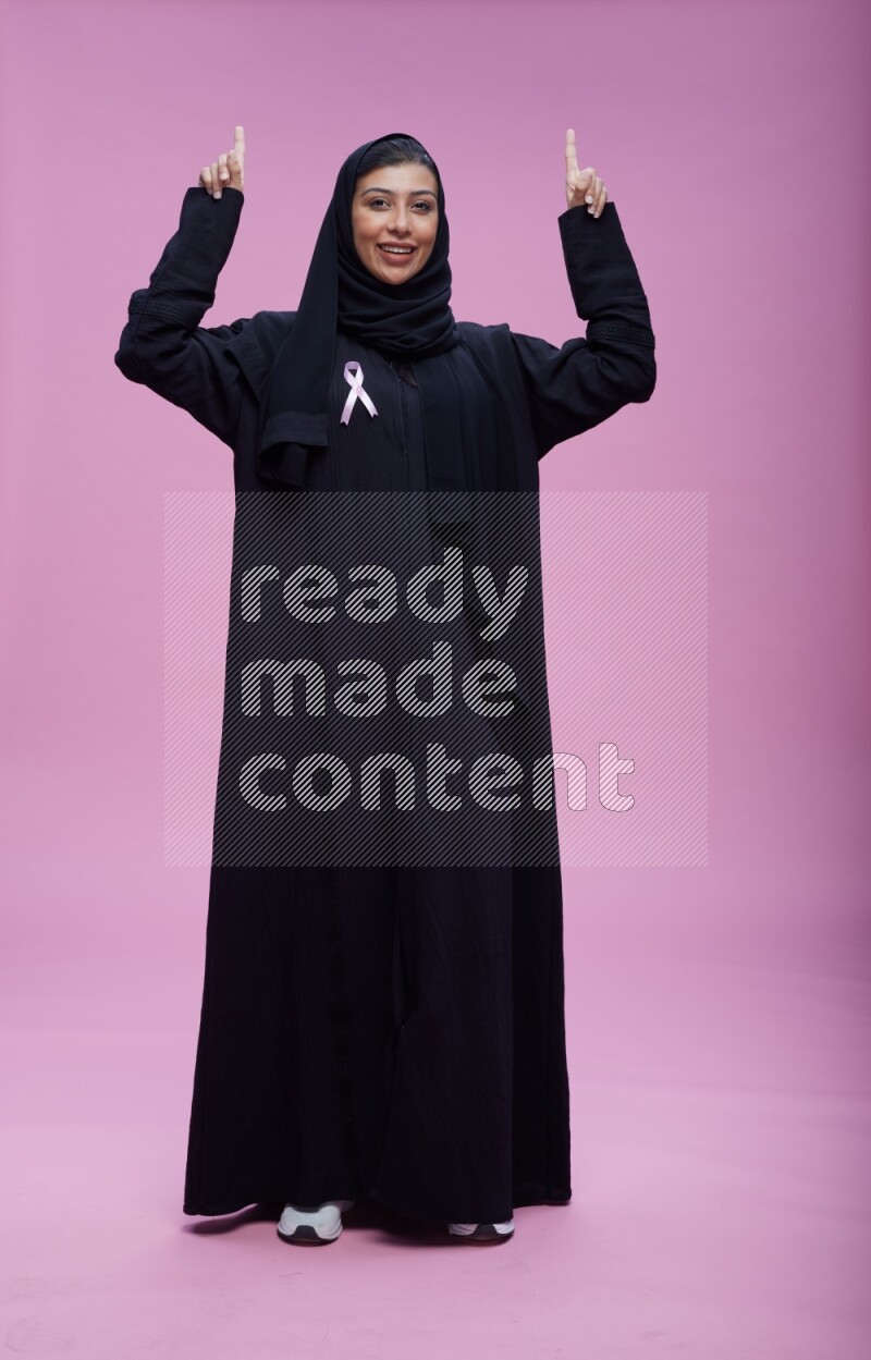 Saudi woman wearing pink ribbon on Abaya standing interacting with the camera on pink background