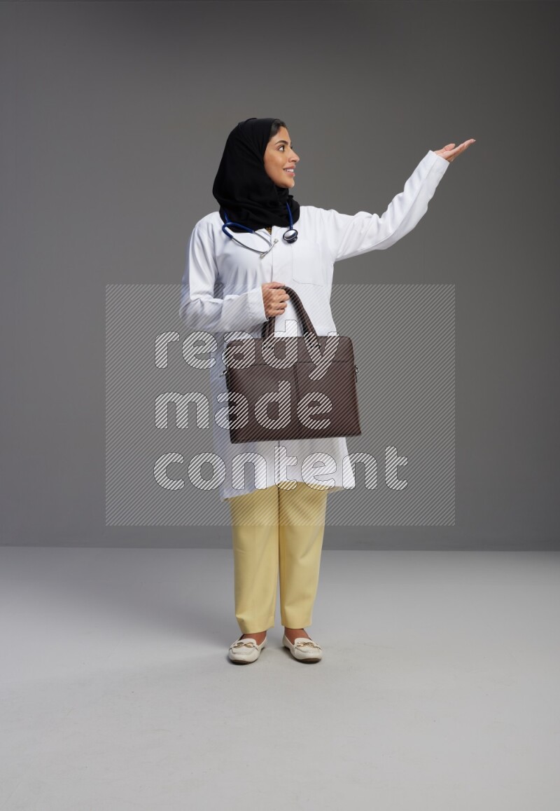 Saudi woman wearing lab coat with stethoscope standing holding bag on Gray background