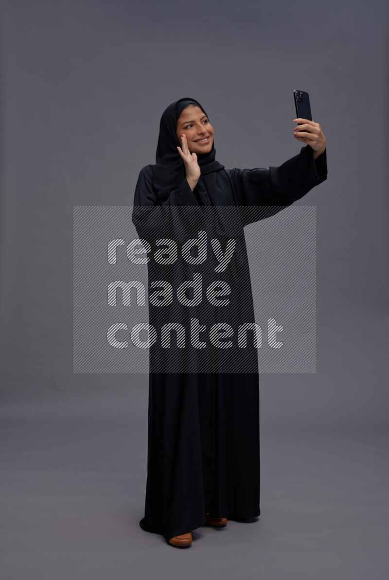 Saudi woman wearing Abaya standing taking selfie on gray background