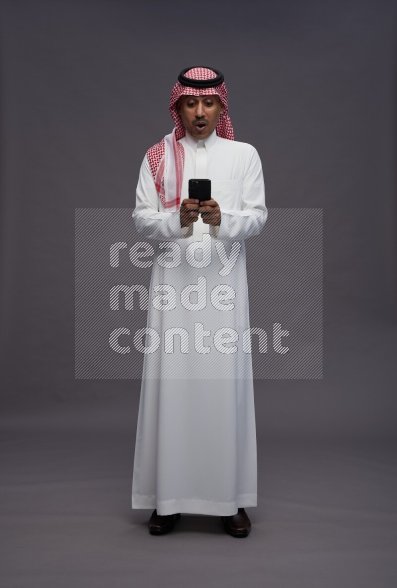 Saudi man wearing thob and shomag standing texting on phone on gray background