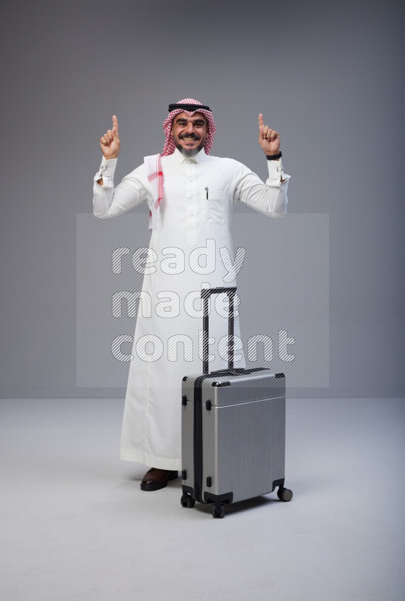 Saudi man wearing Thob and red Shomag standing holding Travel bag on Gray background